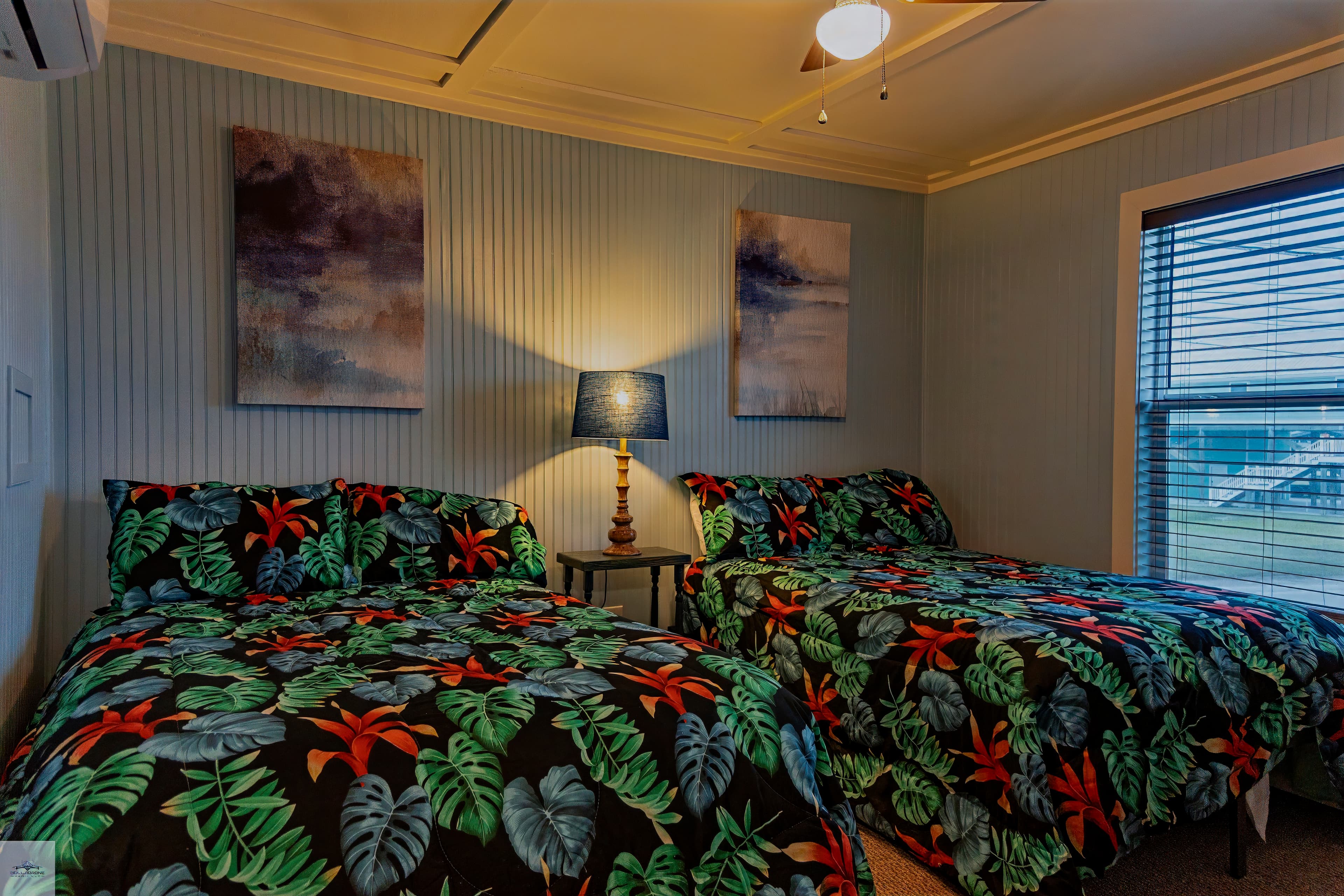 A cozy bedroom with two beds featuring tropical-print bedding, a central nightstand with a lamp, and blue wall paneling.