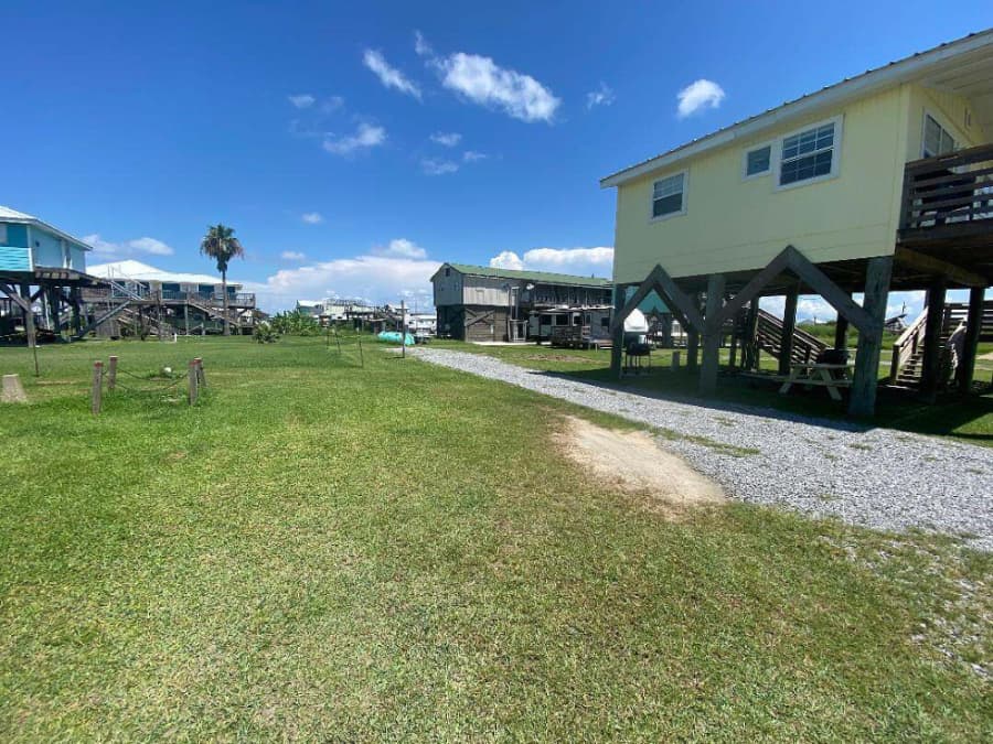 A grassy pathway leads between raised beach houses under a clear blue sky with a few clouds.