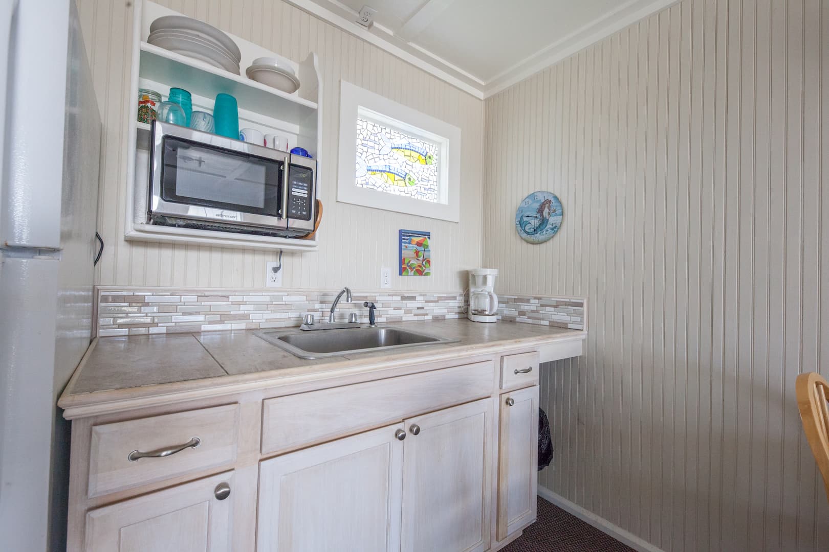 A small, light-colored kitchenette featuring a sink, countertop space, a built-in microwave with shelving, and tile backsplash.