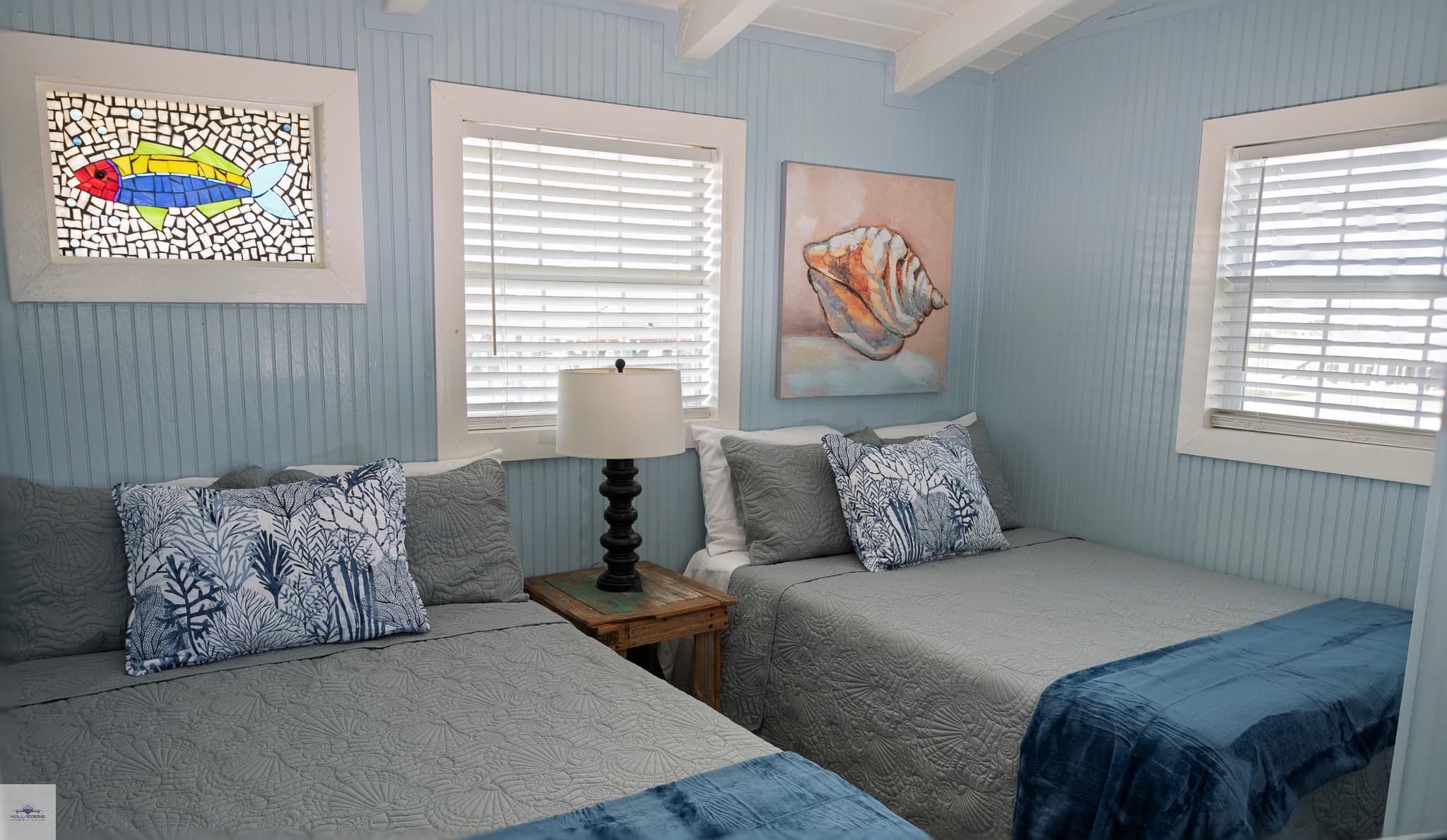 A cozy bedroom featuring two beds with blue accents, a lamp, and coastal-themed decor, with light blue walls and a stained glass window.