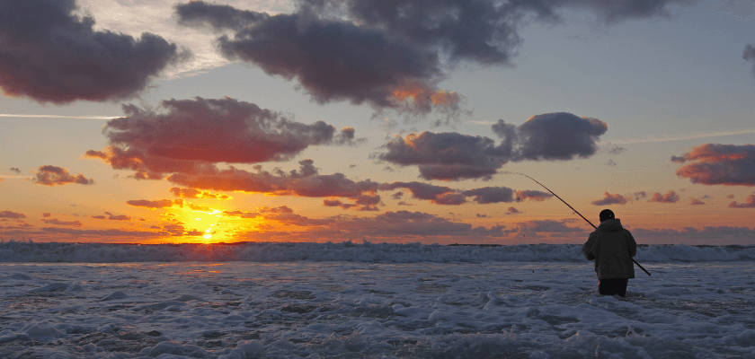 A person stands in the shallow ocean water, fishing at sunset with vibrant clouds overhead.