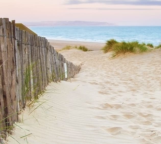 A sandy path bordered by a wooden fence leads to a calm beach and sea under a soft sky.