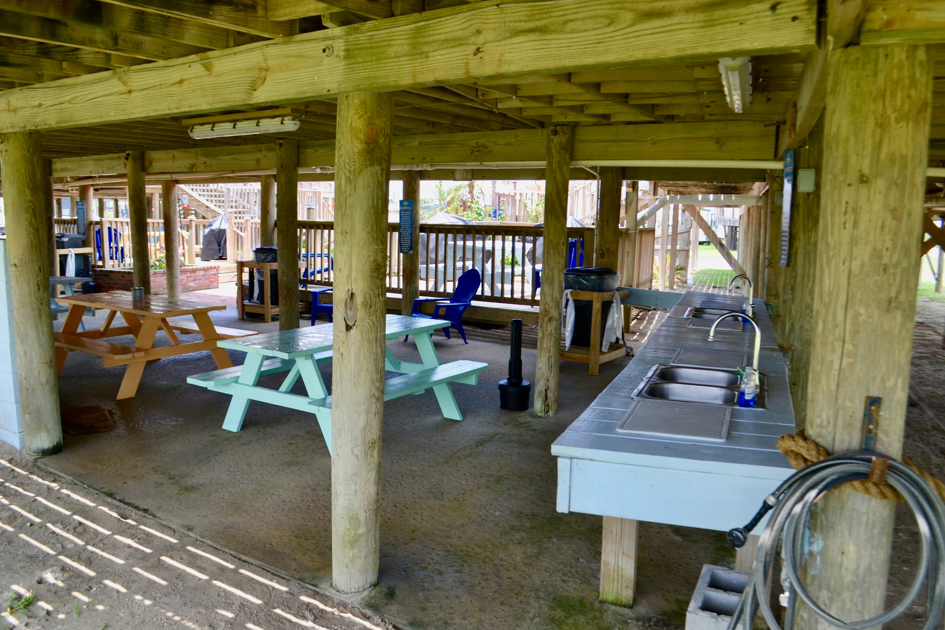 An outdoor covered patio beneath a stilt building, featuring light blue and wooden picnic tables and a long fish cleaning station with sinks.
