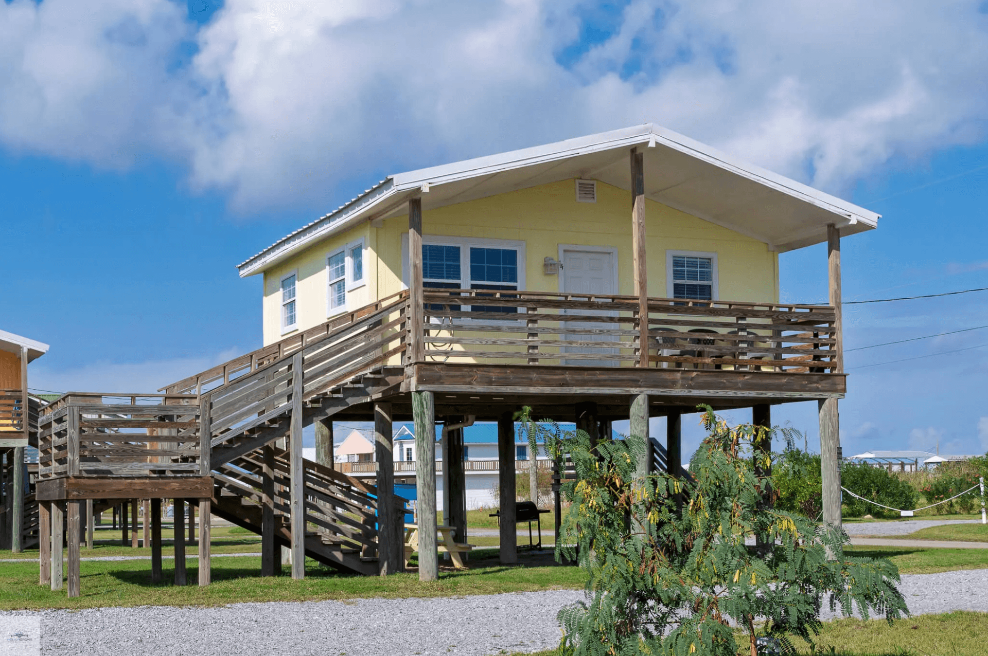 A yellow beach house on stilts with a wraparound deck under a blue sky.