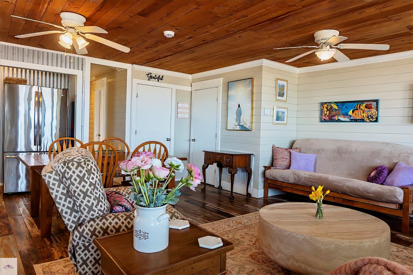 A living room with a wooden ceiling, beige futon, and patterned armchair. A dining table and stainless steel fridge are visible in the background.