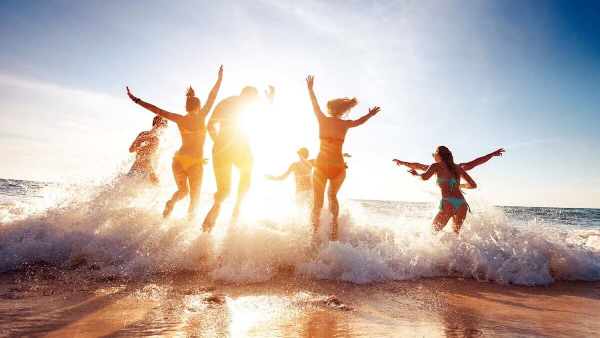 A group of people joyfully splashes in the ocean against a backdrop of a bright sunset.
