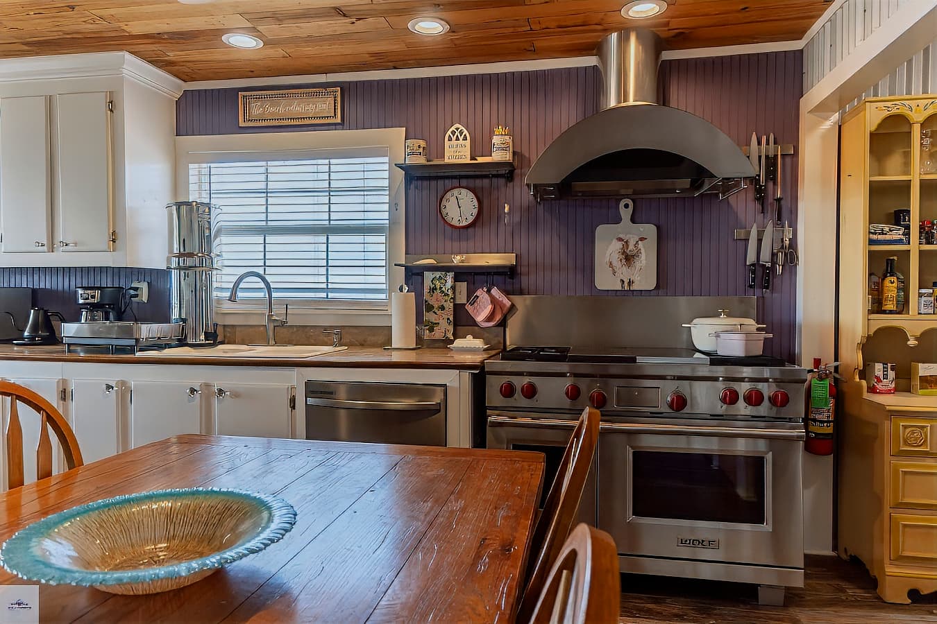 A well-equipped kitchen with white cabinets, a professional stainless steel range and hood, and purple beadboard accent walls.