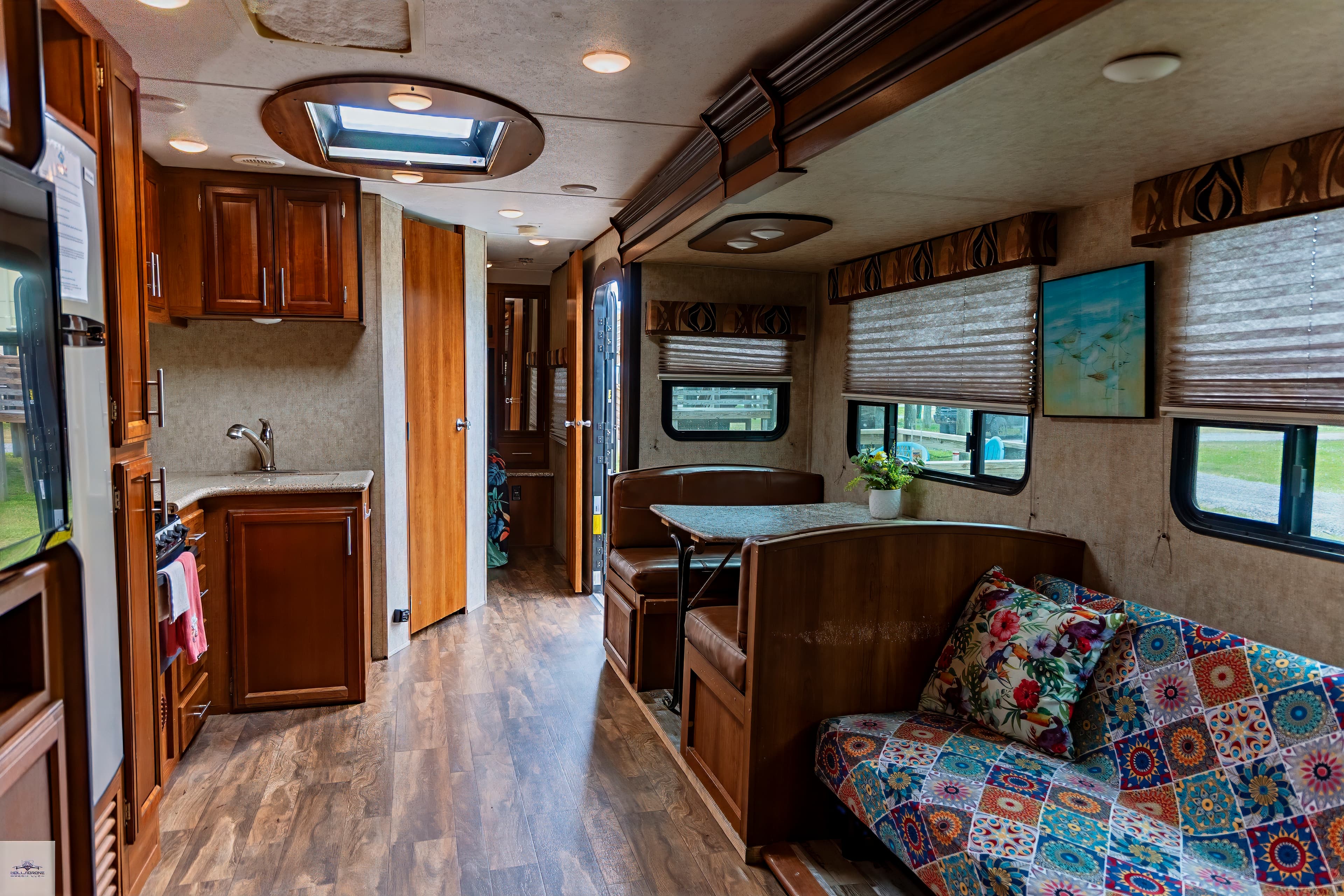 The interior of a camper with wood-look flooring, a kitchenette with dark cabinets, a dinette, and a colorful futon/sofa.