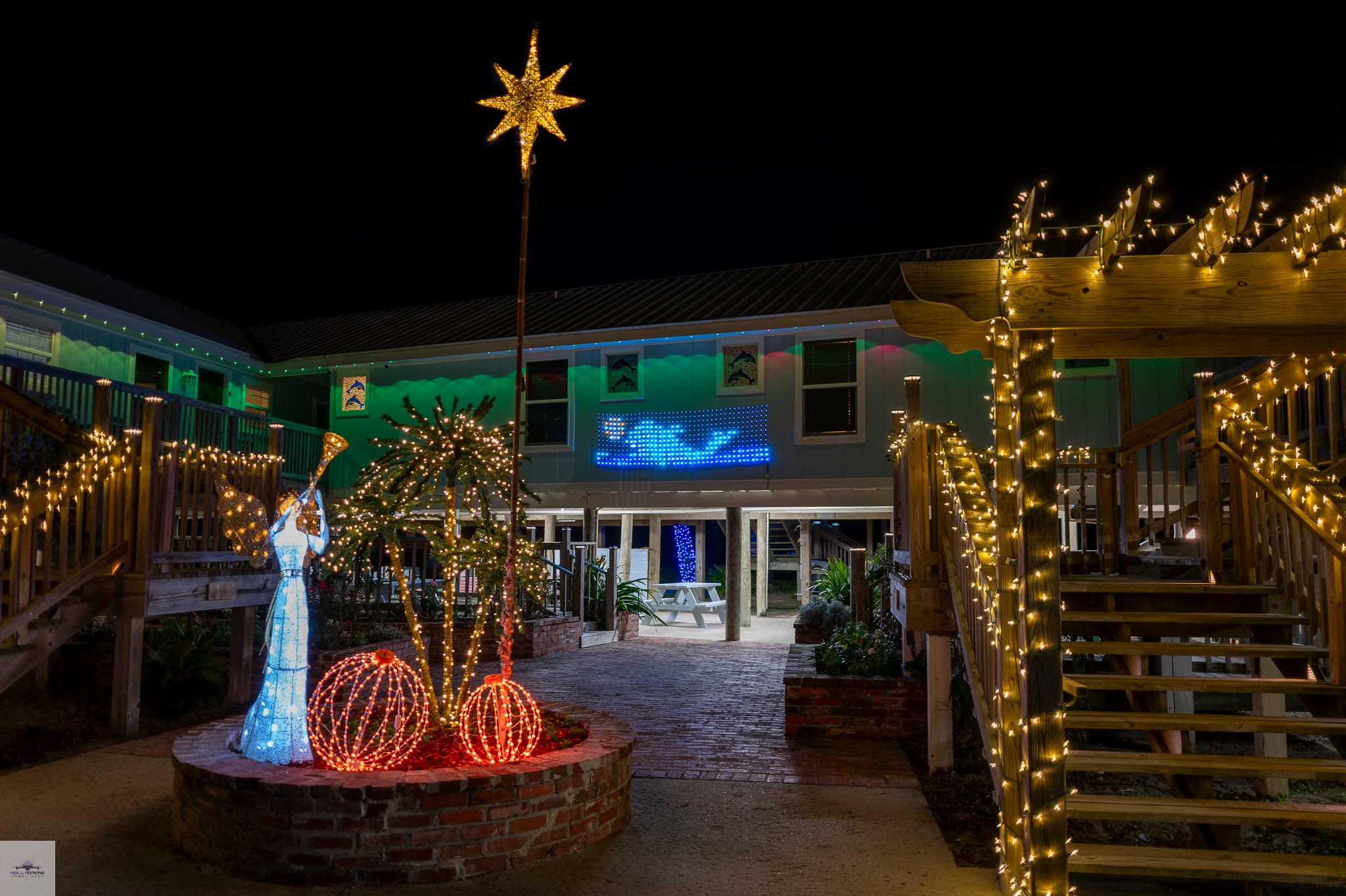 A festive outdoor scene featuring illuminated holiday decorations, including a star, a figure, and glowing spheres, set against a lit building at night.
