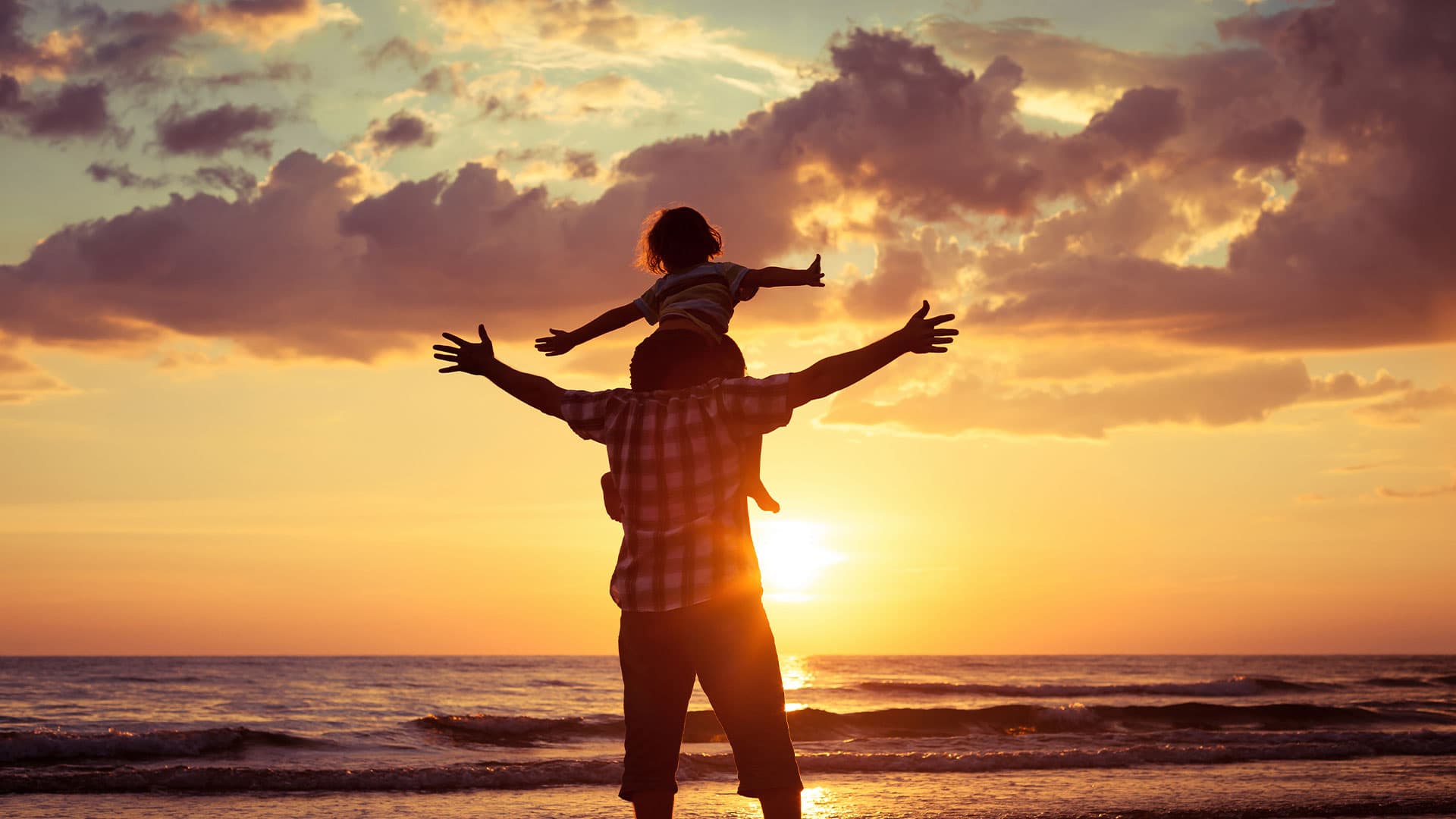 A silhouetted father holds a child on his shoulders against a vibrant sunset at the beach.