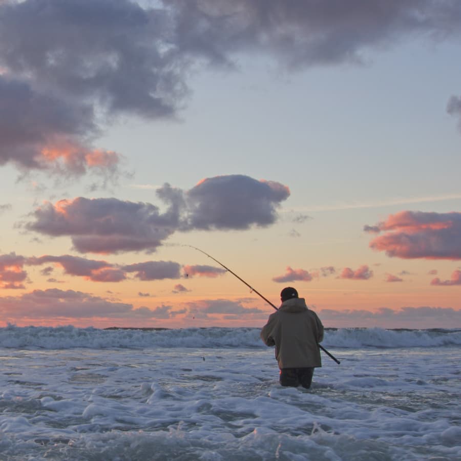 A person fishing in the ocean at sunset with colorful clouds above.