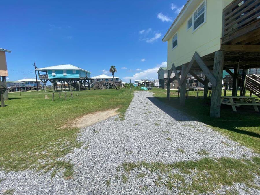 Gravel path between grassy areas and elevated beach houses under a clear blue sky.