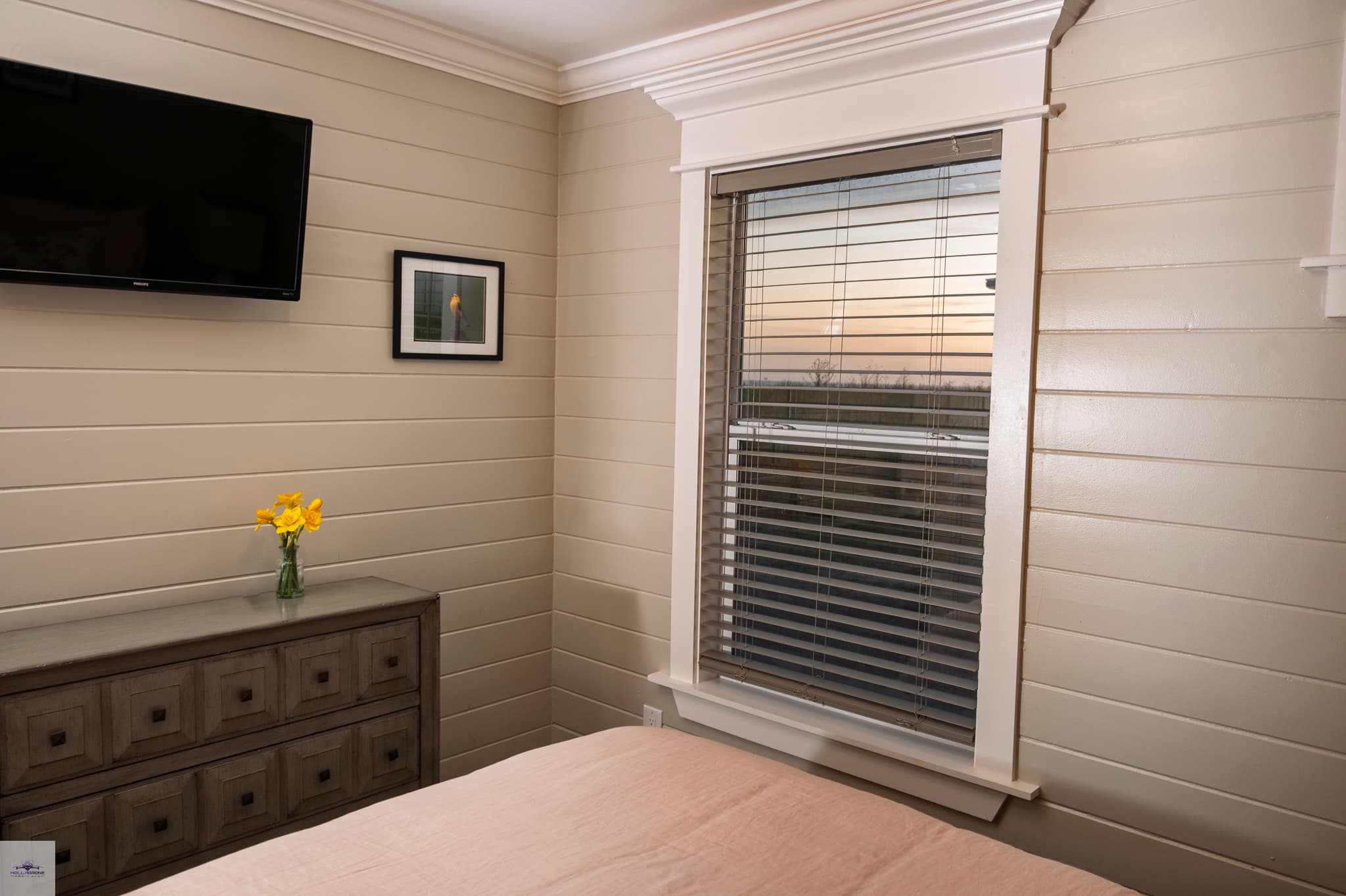 A bedroom with beige walls, a bed with beige bedspread, a chest of drawers, and a TV on the wall.