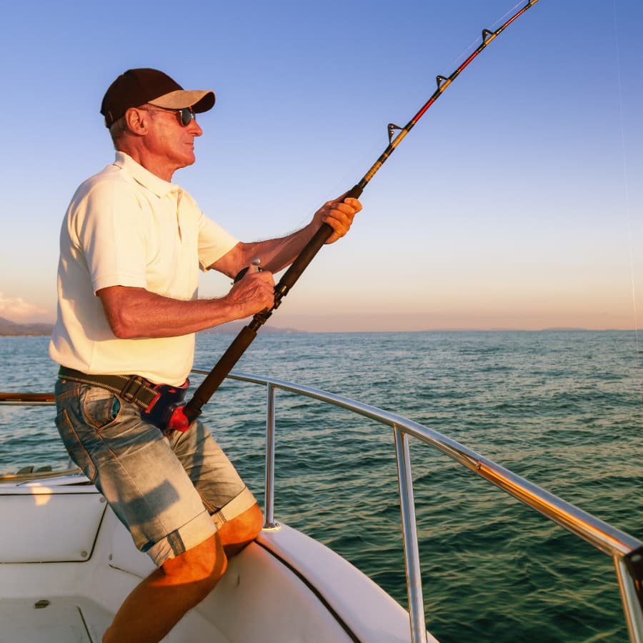 An older man fishing from a boat against a sunset-lit ocean backdrop.