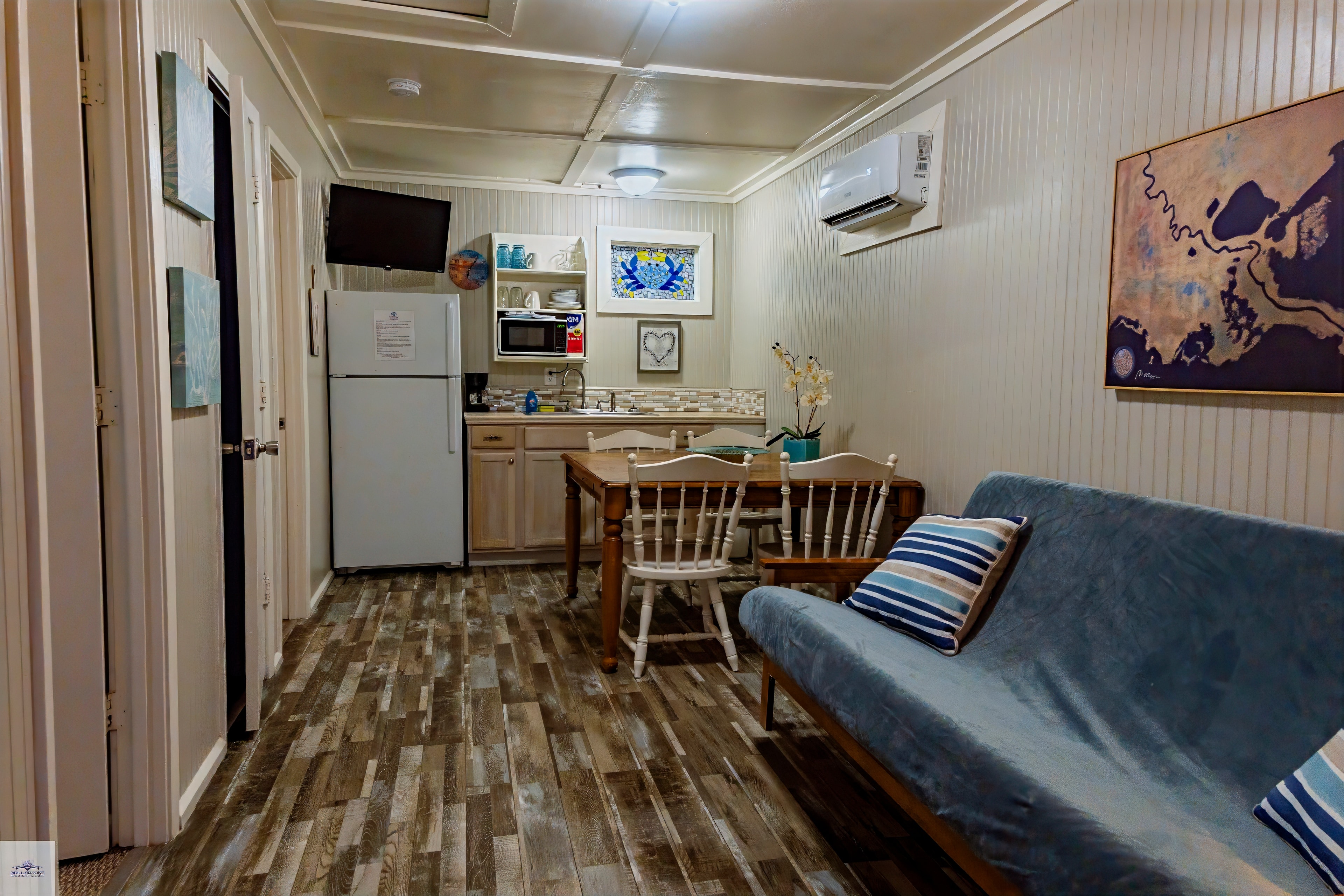 Two bedroom suite entry/living area featuring a blue futon, wooden dining set, a compact kitchenette with a fridge, and coastal artwork.