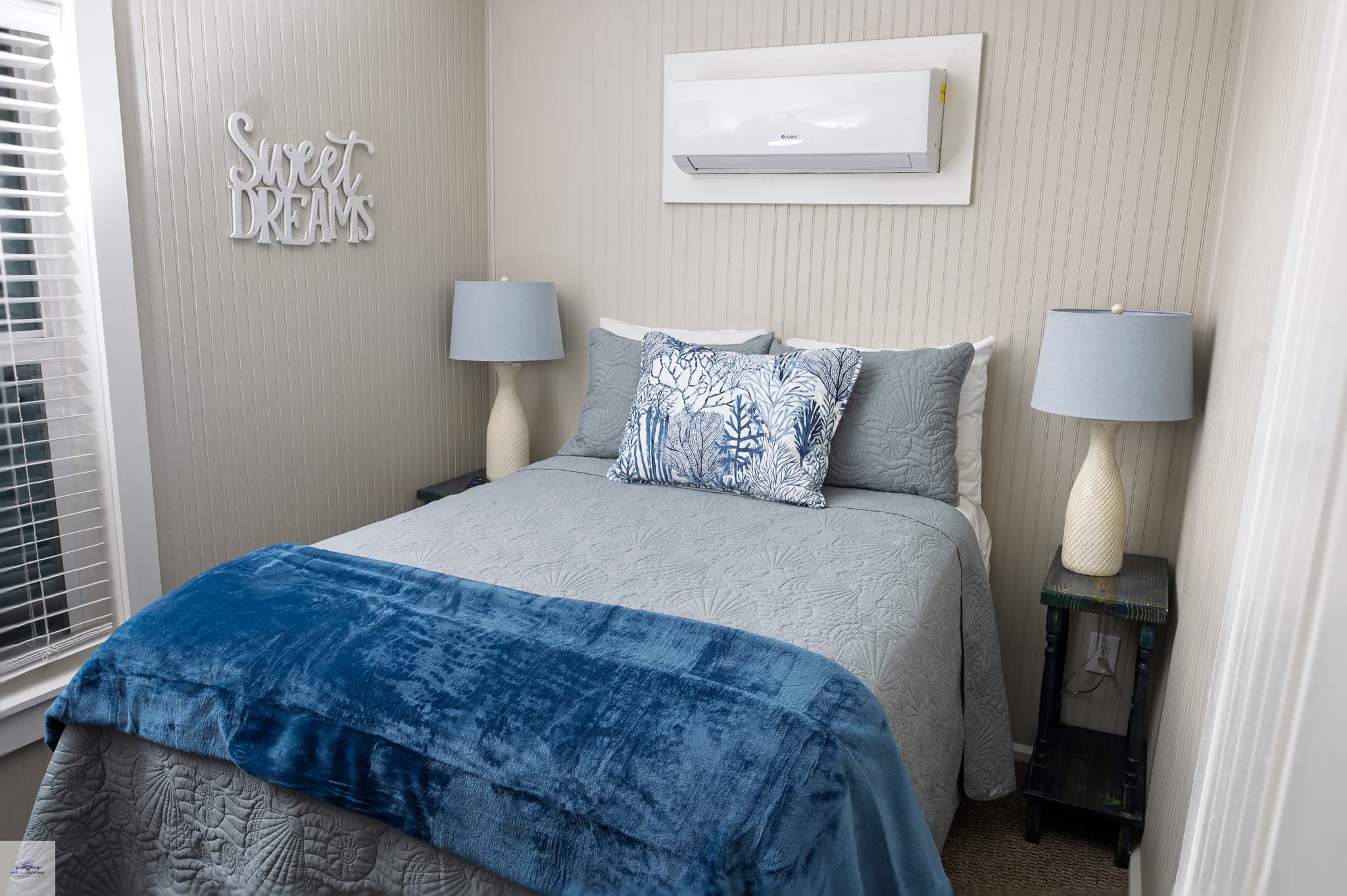 A close-up of a bedroom with a bed featuring a blue and grey bedspread, flanked by two matching lamps, and an AC unit above.