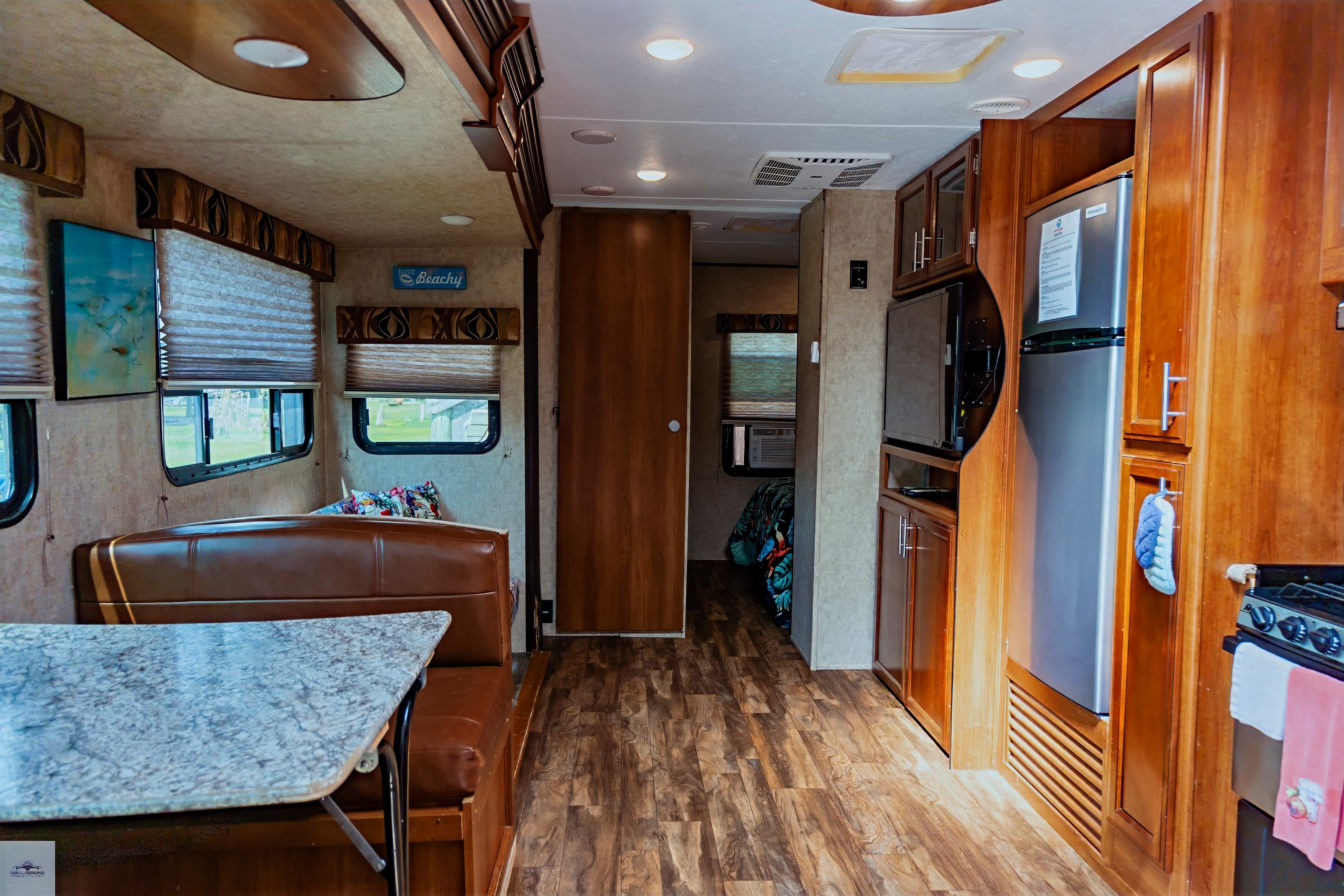 The long interior view of a camper, showing the kitchen with stainless steel appliances, a dinette booth, and wood-look flooring.