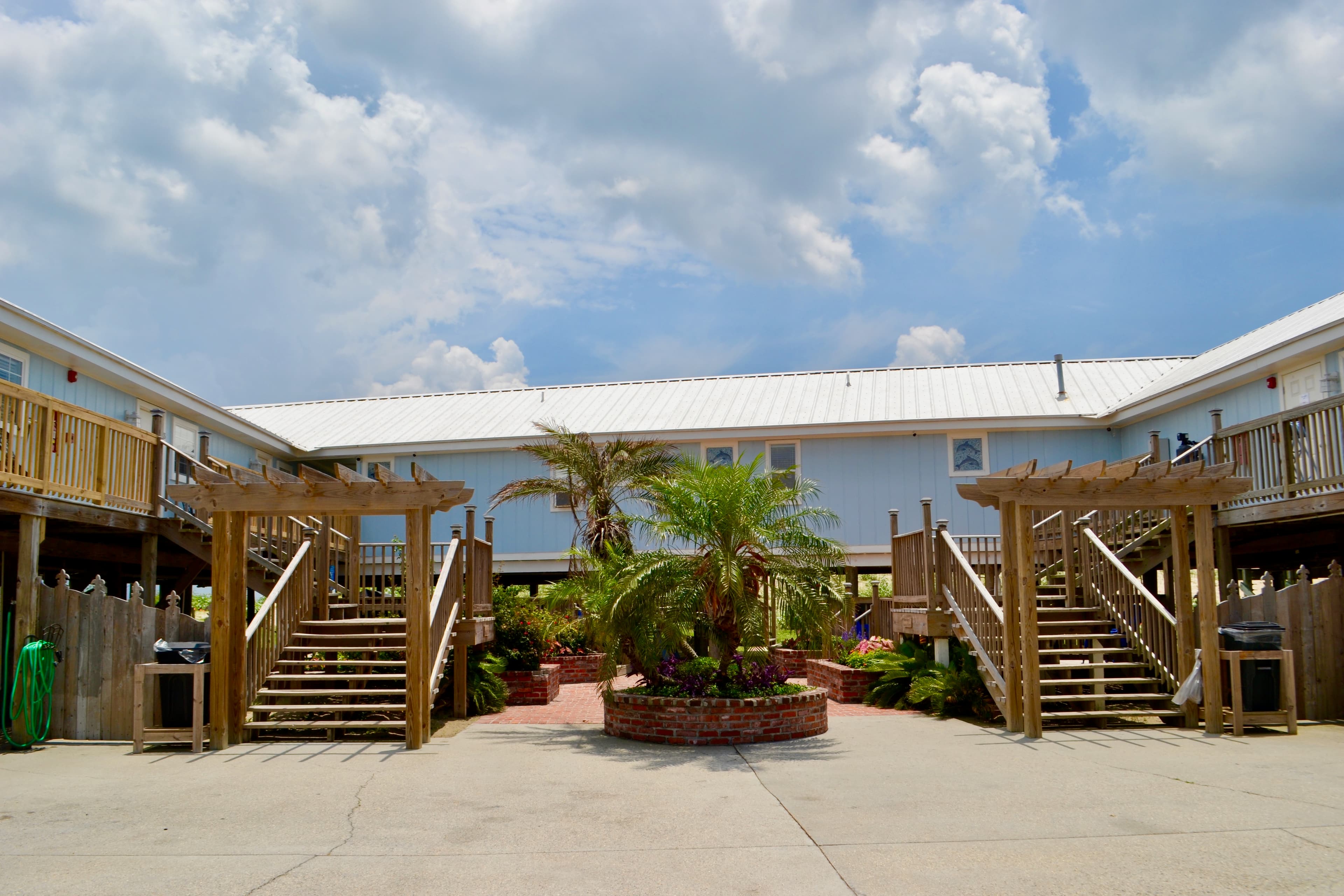 An outdoor view of a courtyard between two rows of light blue stilt buildings. It features a central garden island with palm trees and wooden stairs.