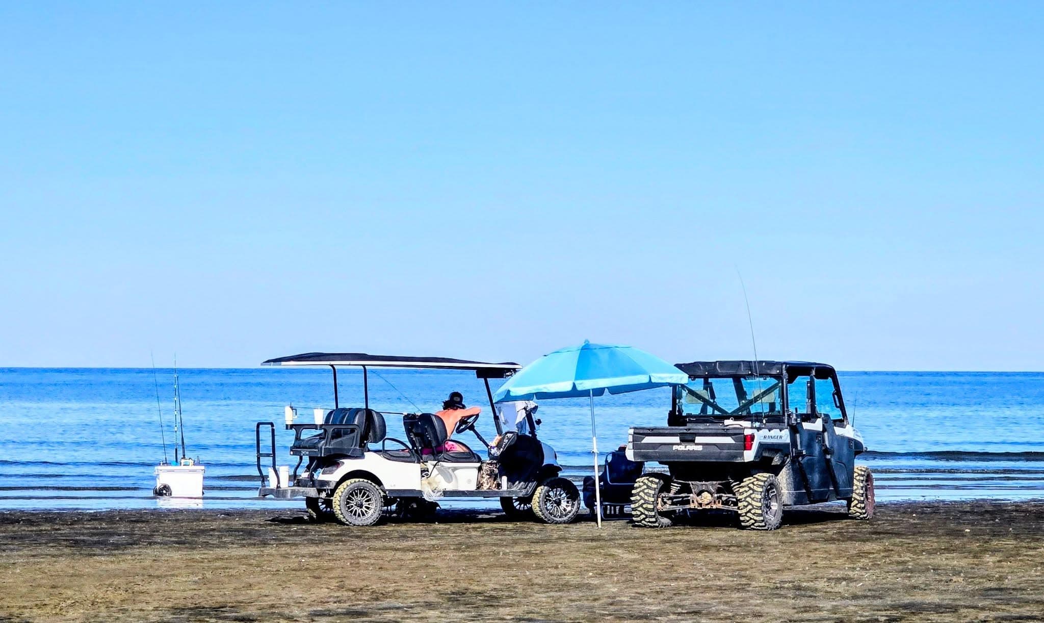 Two off-road vehicles are parked on a beach with a person sitting under an umbrella.